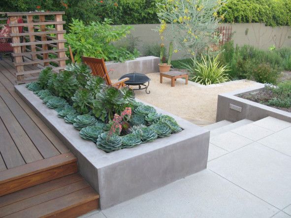 Modern backyard with a rectangular concrete planter of succulents beside a wooden deck, gravel area, and metal fire pit