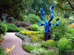 Glossy blue ribbon-like metal sculpture on a pedestal beside a curved brick path and yellow garden flowers