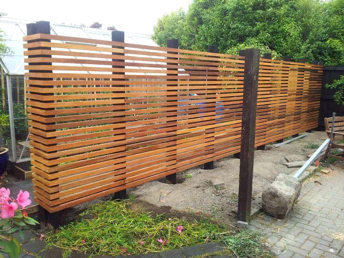 Modern horizontal-slat wooden privacy screen on dark posts beside a glass greenhouse and paved walkway