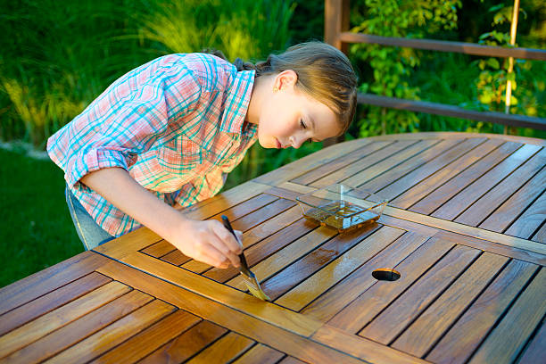 Person brushing clear wood finish onto a slatted outdoor wooden table in a sunny garden