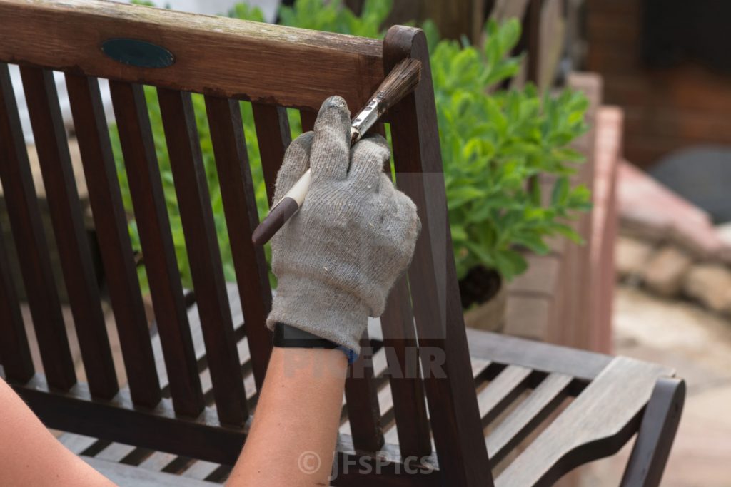 Gloved hand applying dark brown stain with a paintbrush to vertical slats of a wooden garden chair