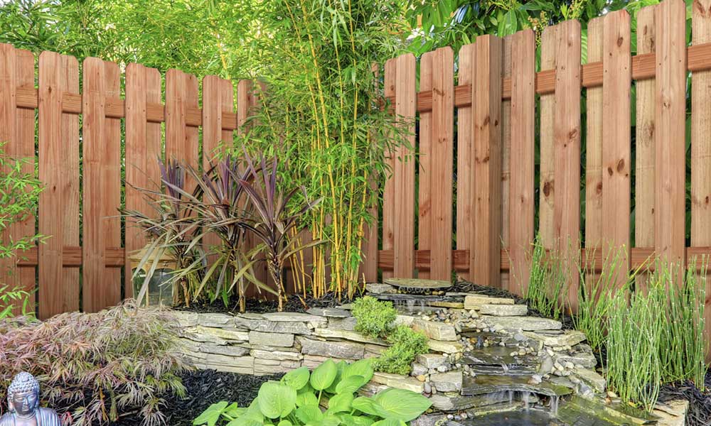 Wooden privacy fence behind a stacked-stone waterfall, bamboo, hostas, and ornamental plants in a backyard corner