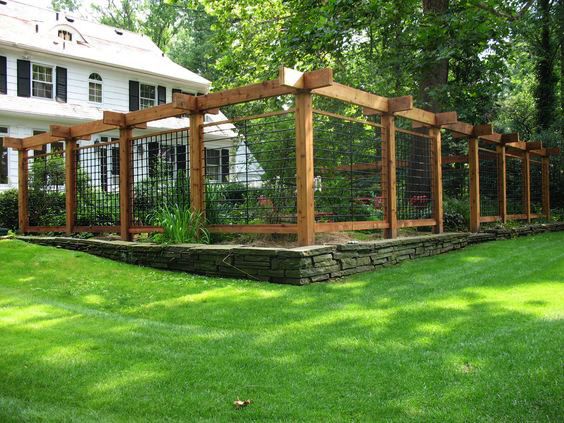Timber pergola-style fence with black wire-mesh panels on a low stone wall in front of a white two-story house