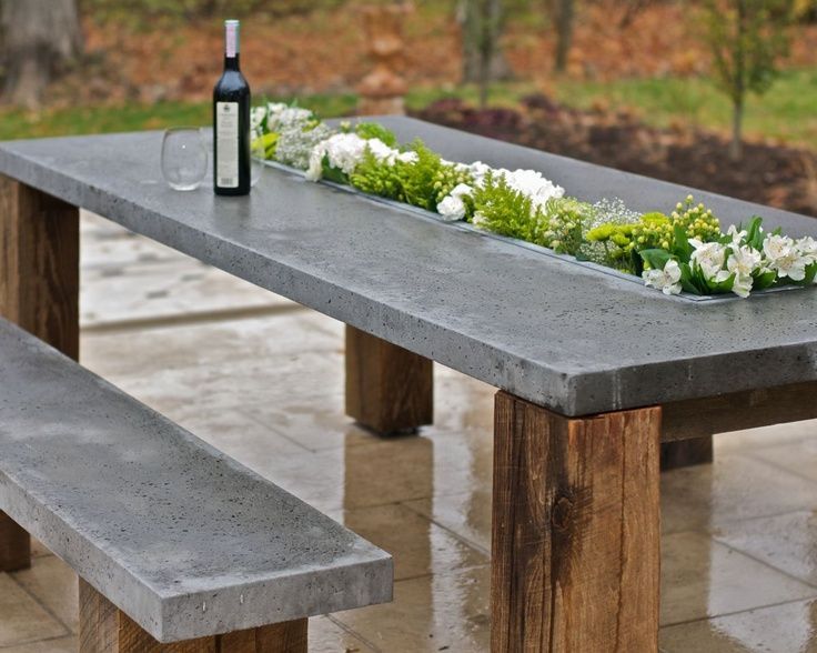Concrete-top outdoor dining table with built-in planter trough of white flowers and rustic wood legs on a stone patio