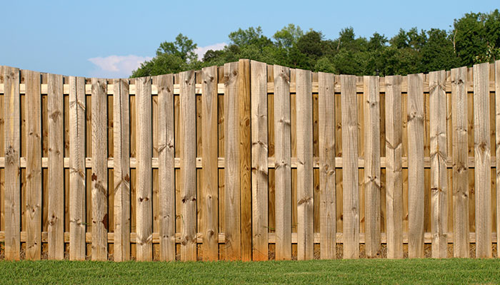 Weathered light-brown wooden privacy fence with scalloped dog-ear pickets above a green lawn